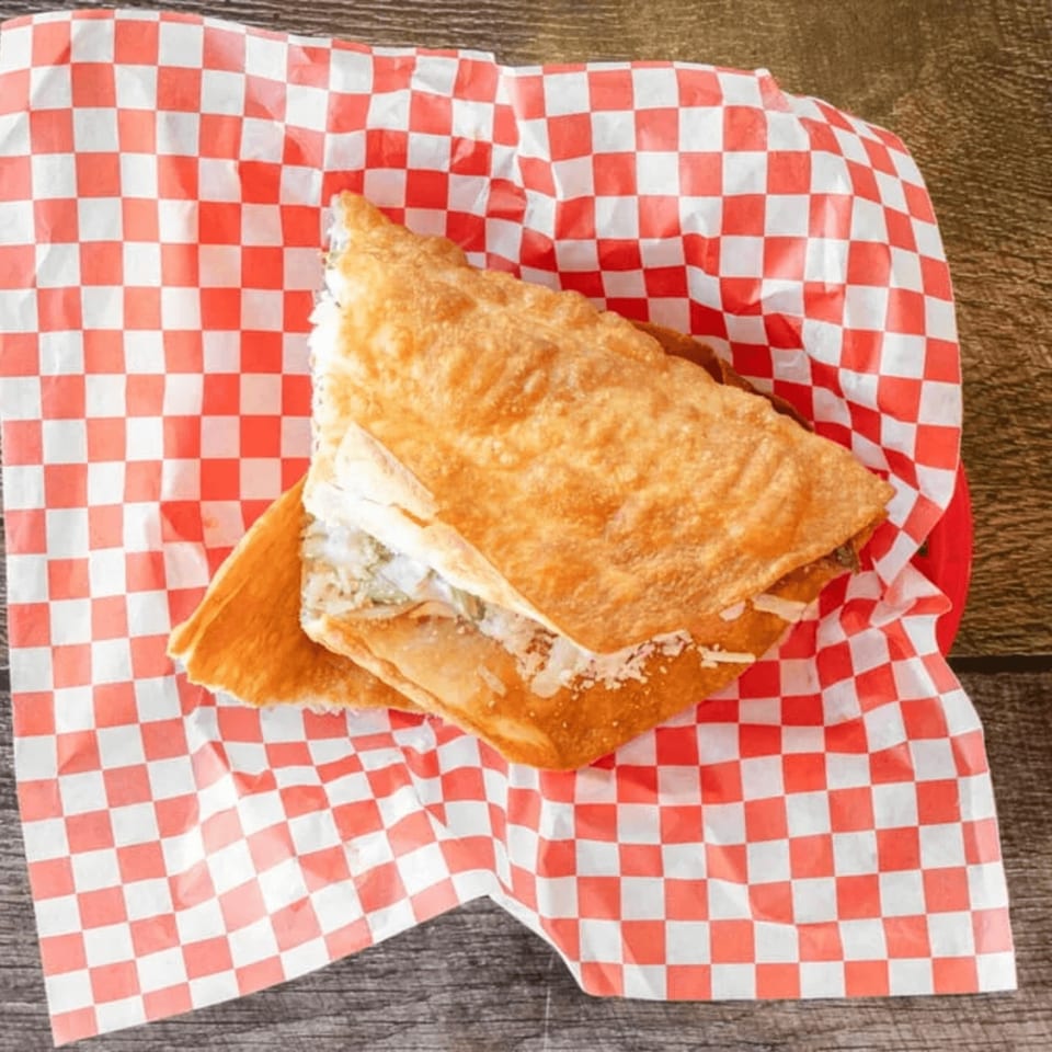 A golden-brown fried quesadilla, filled with cheese and meat, rests on red and white checkered paper.