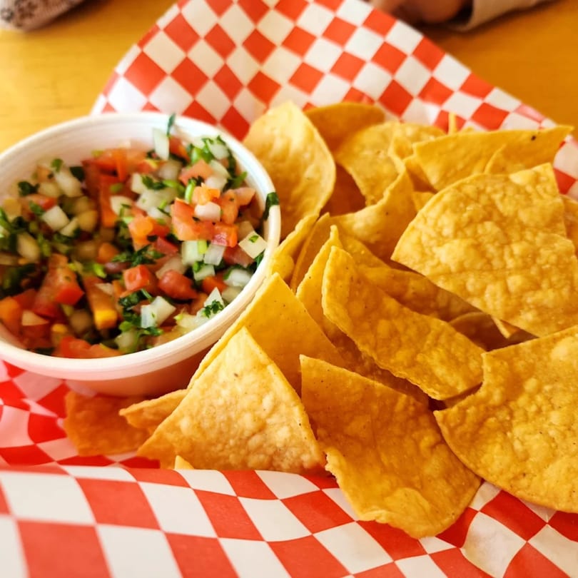 A white bowl of fresh pico de gallo next to a pile of tortilla chips on red and white checkered paper.