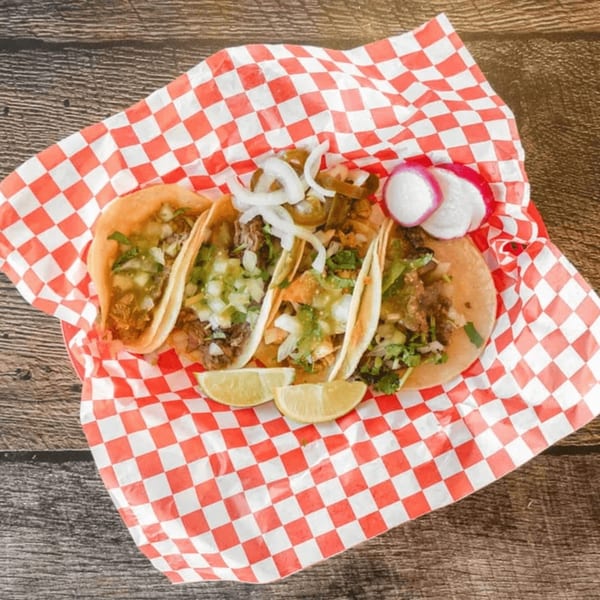 Four Mexican street tacos with meat, onions, cilantro, limes, and radishes on checkered paper.